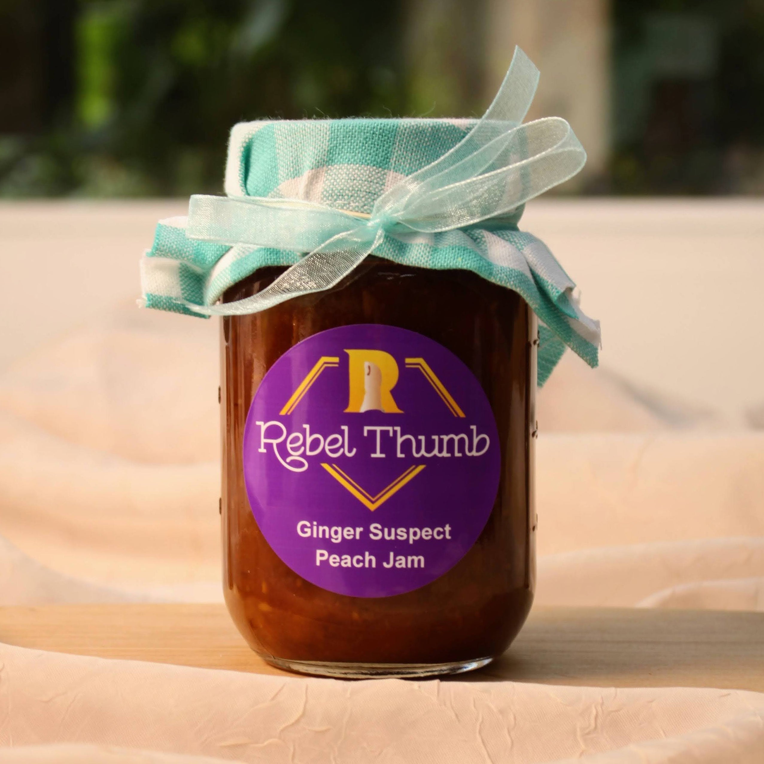 California made jar of Rebel Thumb organic Ginger Suspect Peach Jam with a turquoise ribbon on a beige surface.